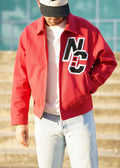 A man wearing a red zip-up jacket with a large black and white 'NC' patch on the chest, a white t-shirt, light blue jeans, and a black and white baseball cap, standing outdoors in front of concrete steps.